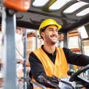 Young man operating a forklift loader in distribution warehouse. Male worker in hardhat and reflective vest driving a forklift.
